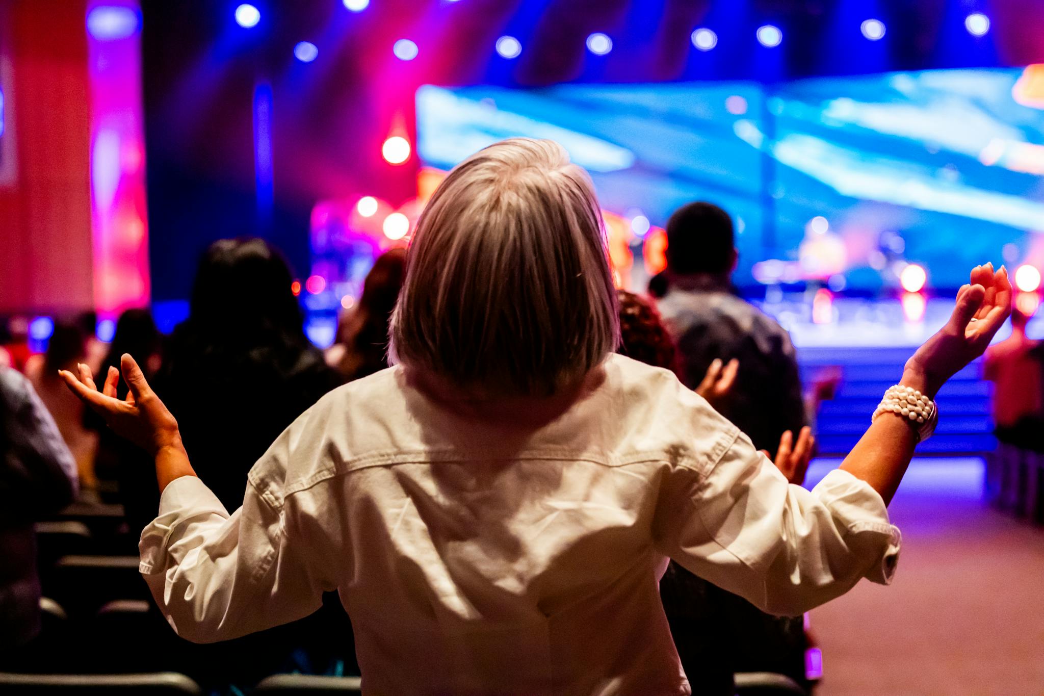 A vibrant indoor worship service with people raising hands in praise and colorful stage lighting.