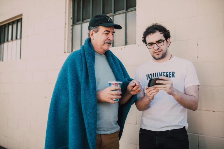 A volunteer helping a homeless man with smartphone services outdoors.
